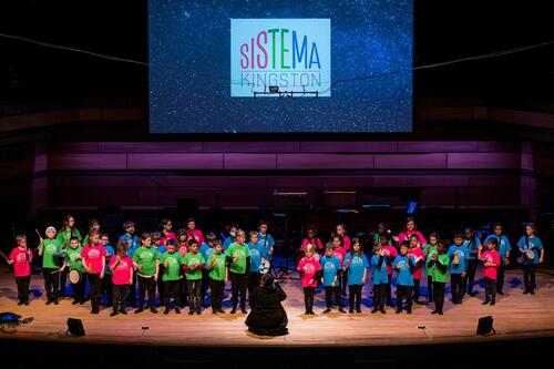 Sistema Kingston students stand on stage in rows at a performance.