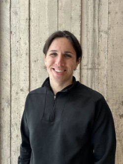 Tristan smiles in front of a stone wall in a black quarter zip sweater.