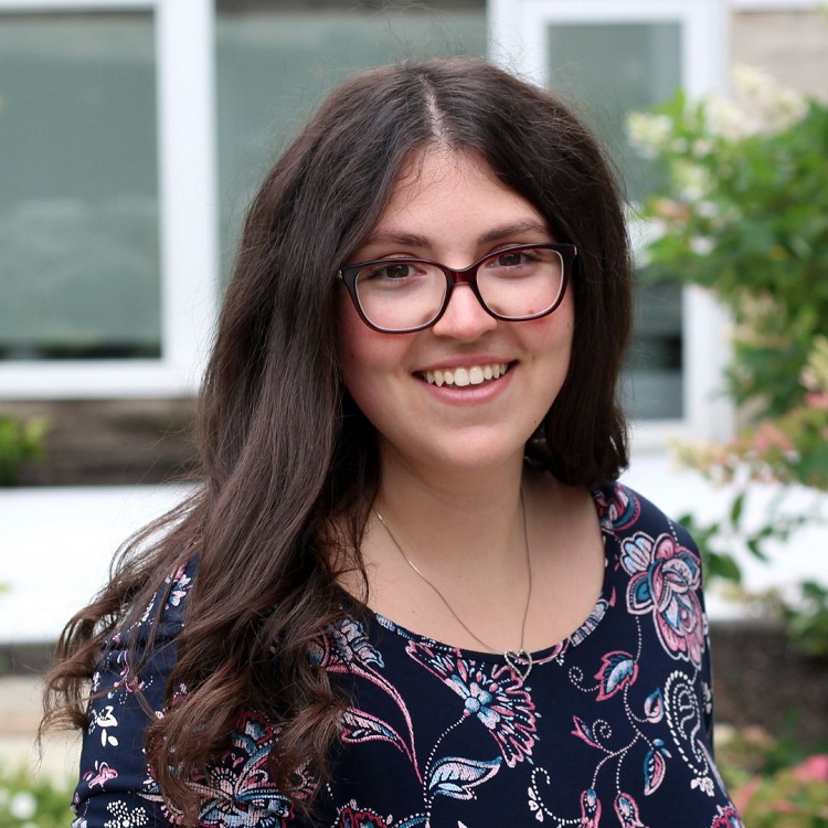 Alexandra Bosco smiles outside, she wears glasses and a purple floral shirt.