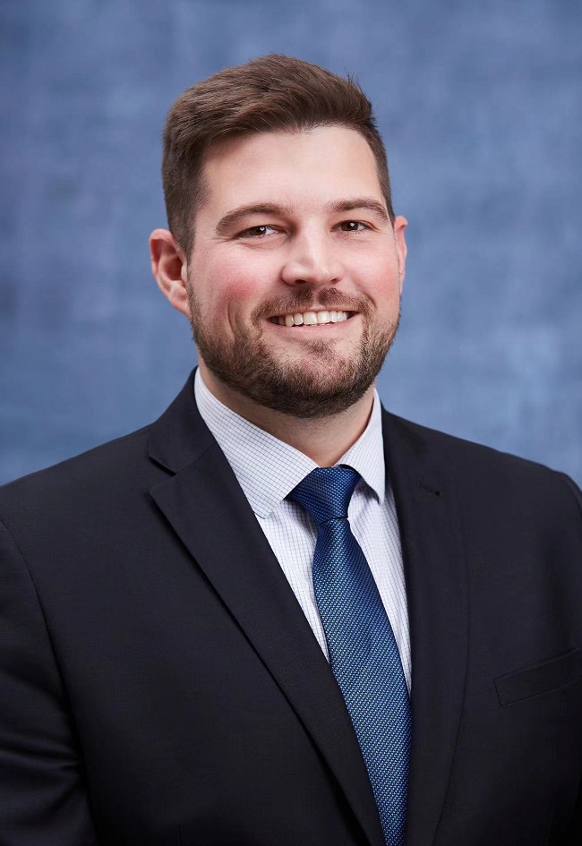 Ryan Parry smiles against a blue background wearing a collared shirt.