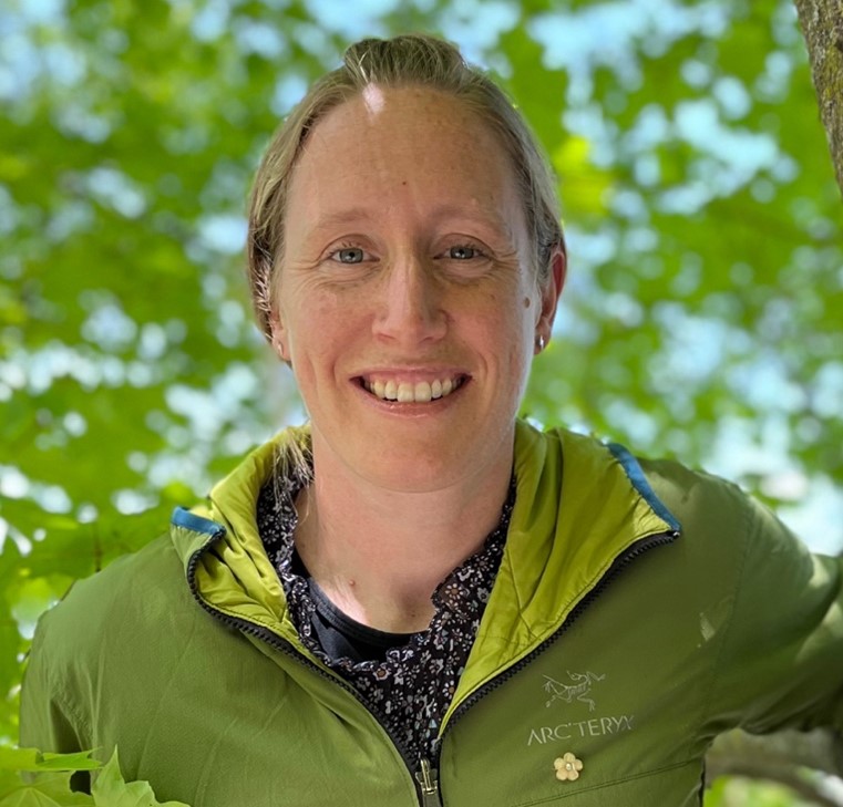 A woman with blond hair and blue eyes smiles in a green coat that matches green leaves around her