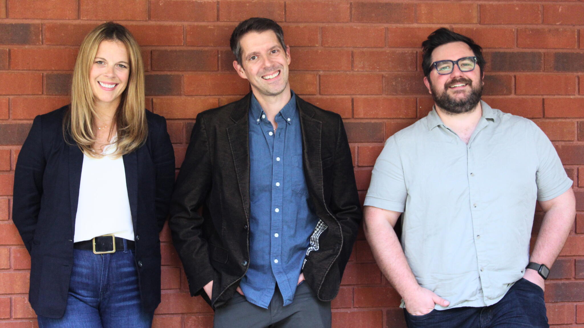 Launa Gauthier, Jason Shulha, and Nate Axcell smile standing against a red brick wall.