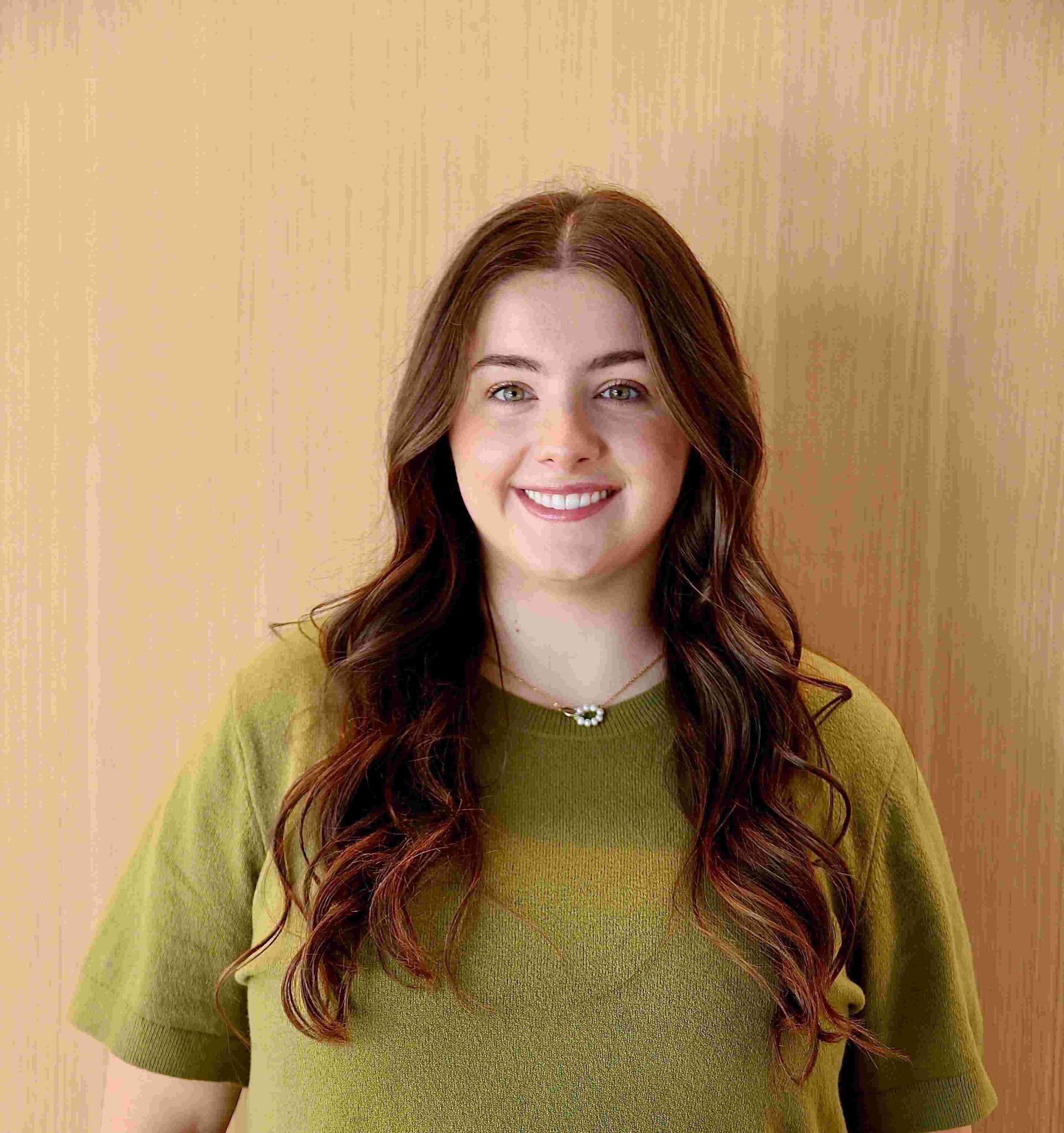 A woman smiling in front of a wood wall with a green sweater on
