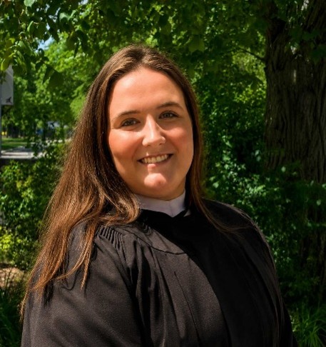 A woman with long dark hair smiles in a black graduate gown