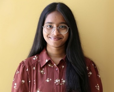 A woman with glasses and dark hair smiles in front of a gold wall 