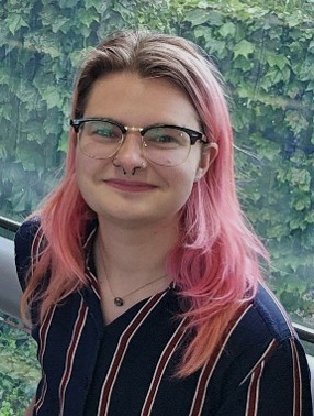 A person with glasses and pink hair smiles at the camera