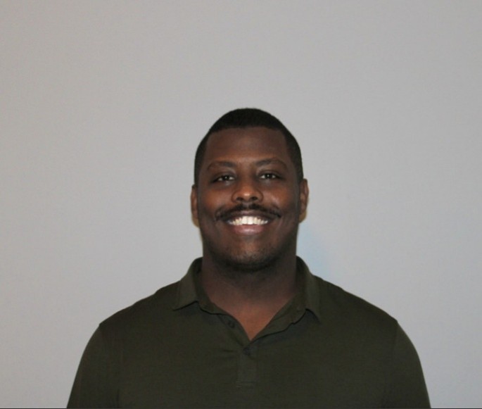 A black man smiles at the camera in front of a white wall