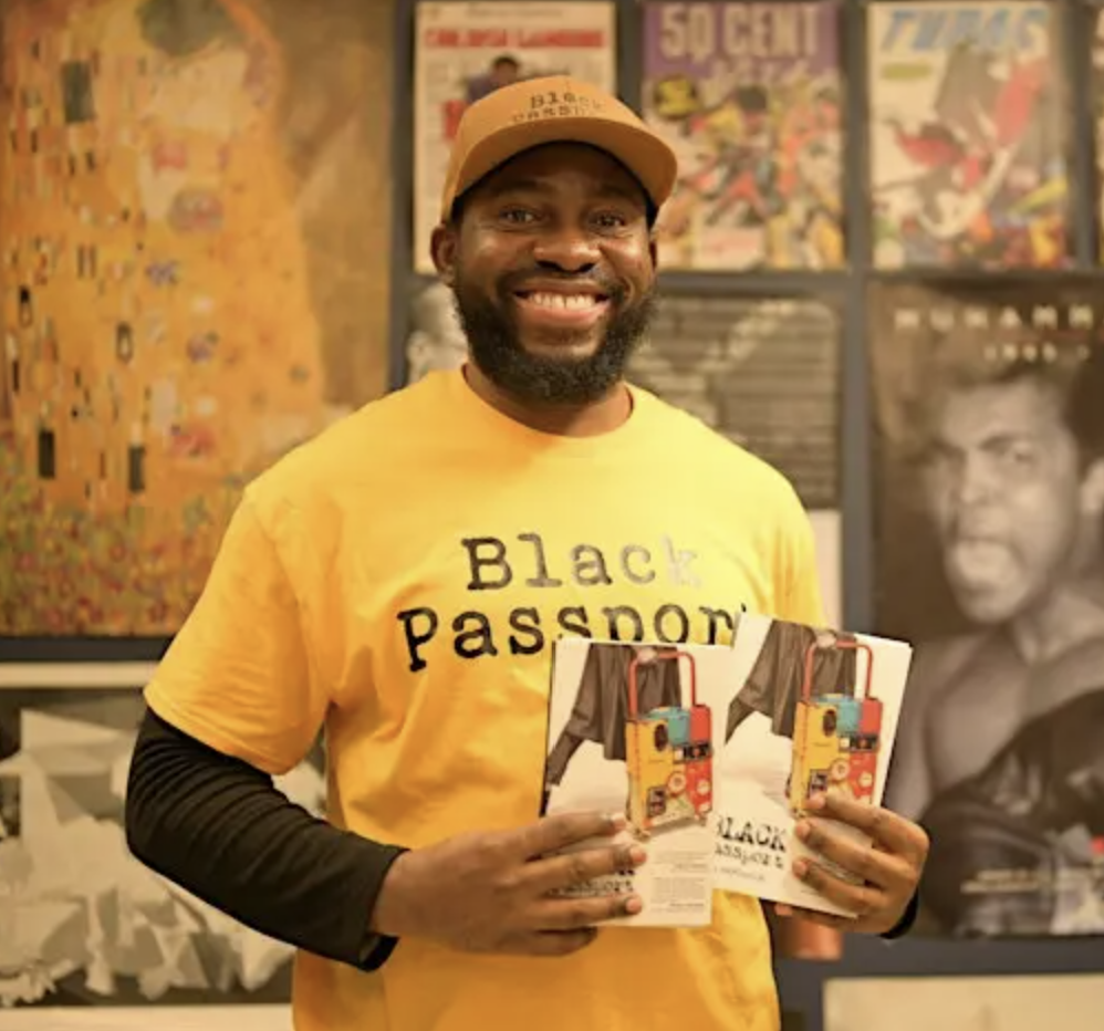 Paul Akpomuje smiles holding two copies of his book Black Passport. He wears a yellow t-shirt that reads "Black Passport" in black writing.