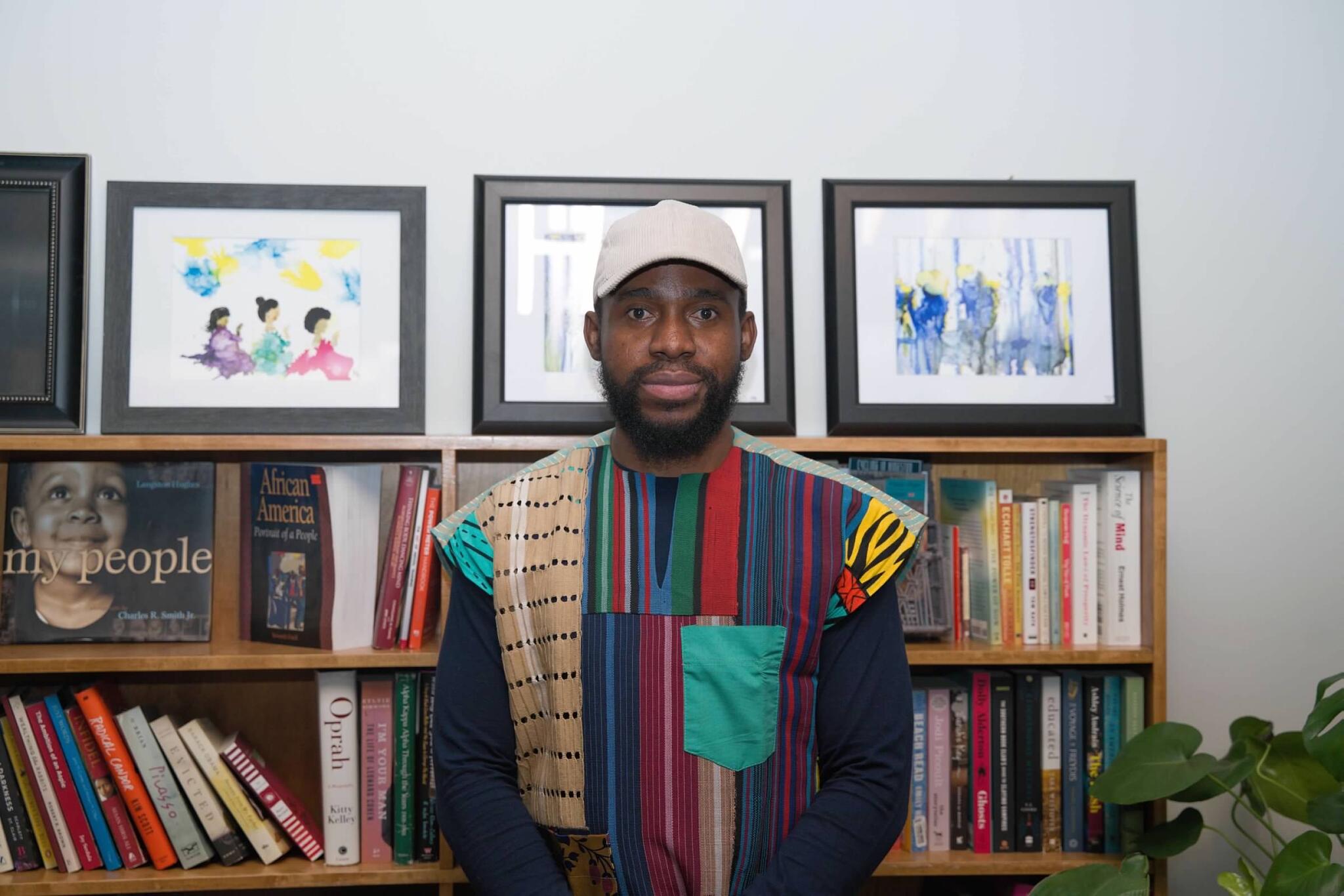 Paul stand in front of a bookshelf in a white baseball hat