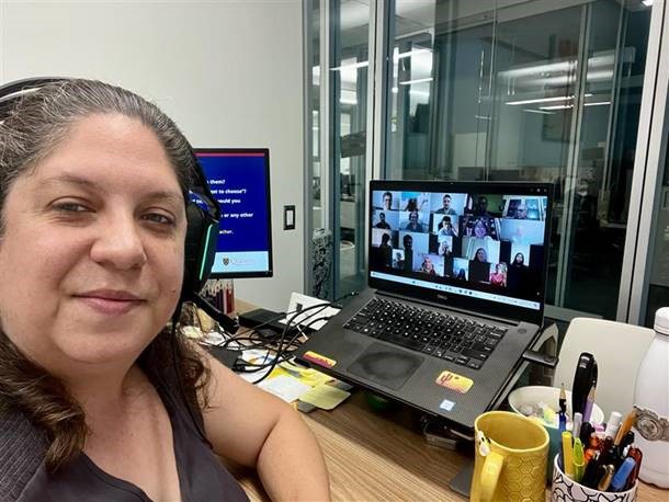 A woman looks at the camera with a computer in the background that shows many students participating in online learning