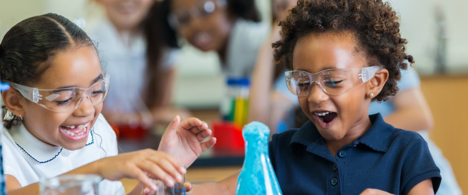 Two young black children giggle while performing a science experiment and wearing safety goggles