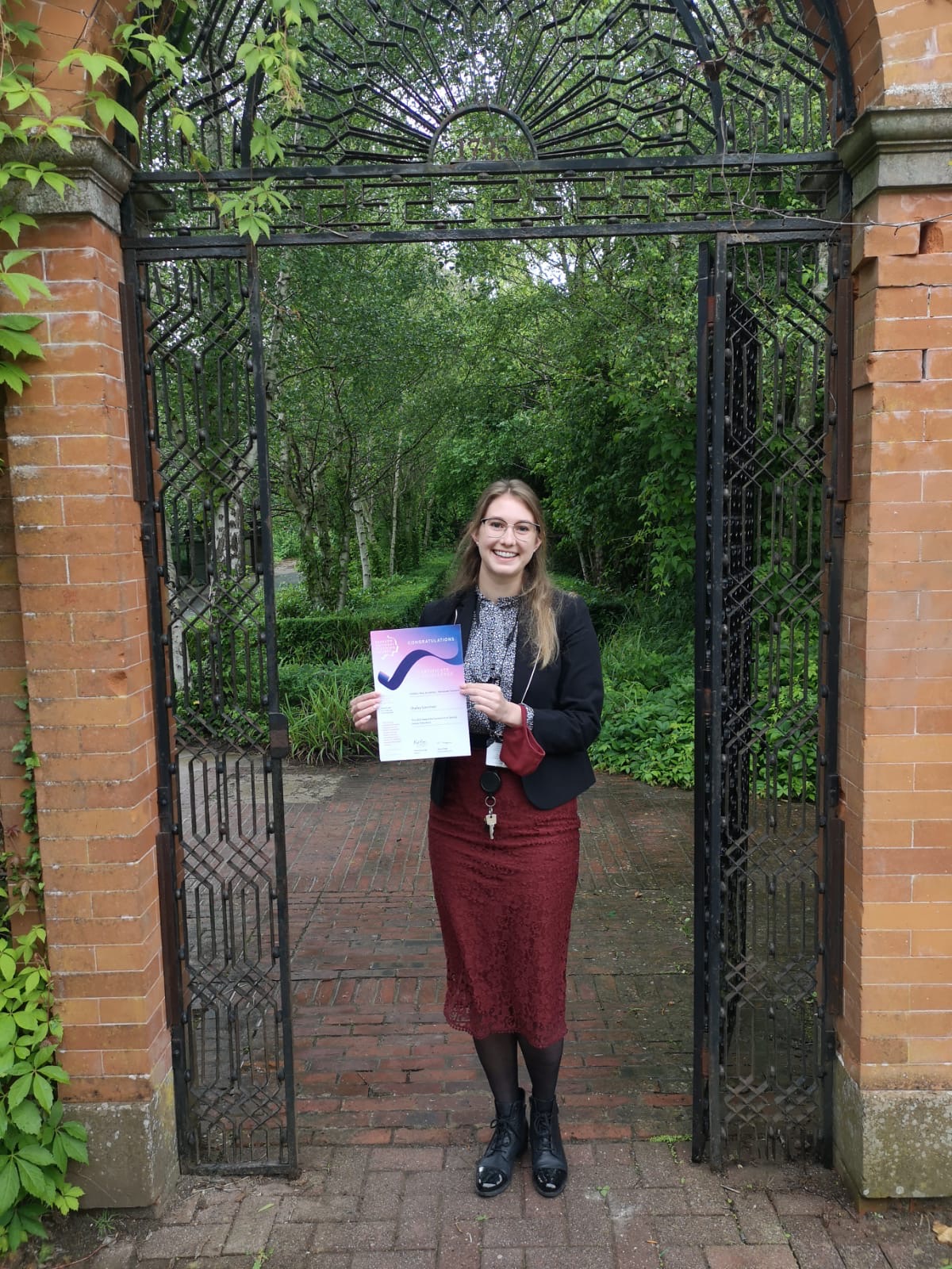 A woman stands underneath a brick and iron archway holding a certificate