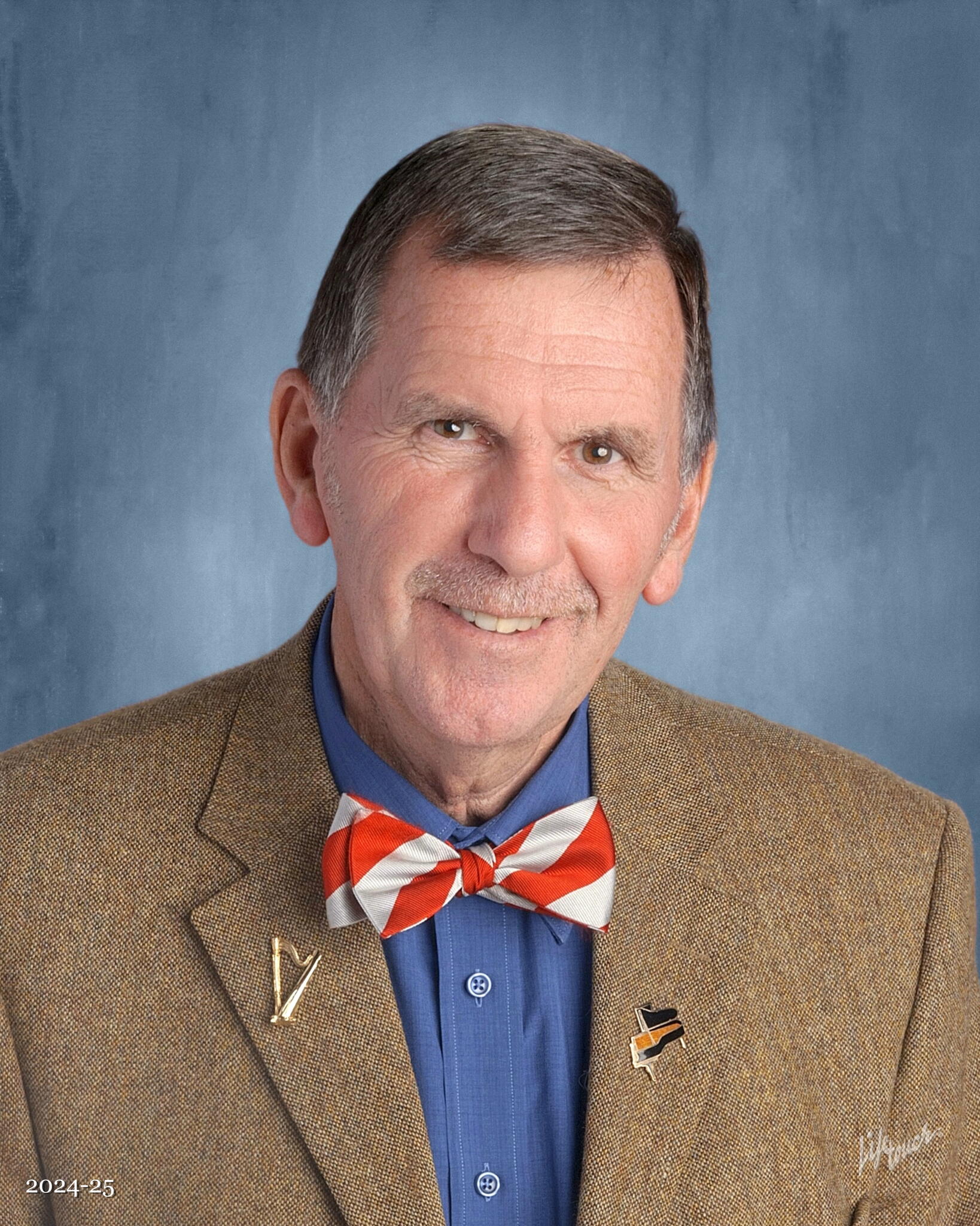 James Deagle smiles wearing a blue collared shirt, striped bowtie, and brown blazer.