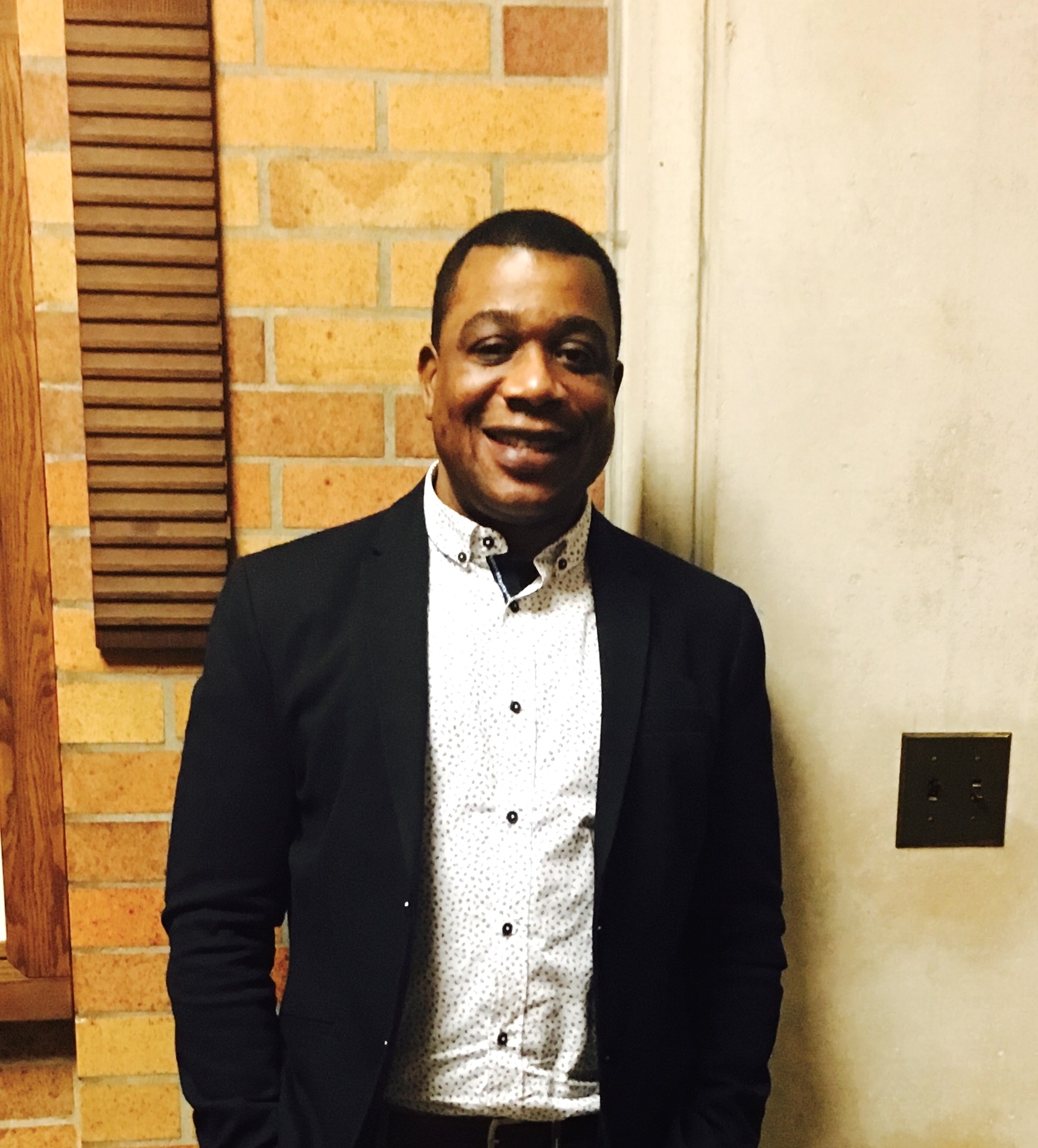 Leroy Baker smiles standing against a brick wall wearing a collared shirt and blazer.