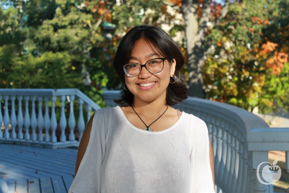 Giselle Castillejos smiles in a white knit sweater and glasses standing outside in front of a curved wooden railing and trees.
