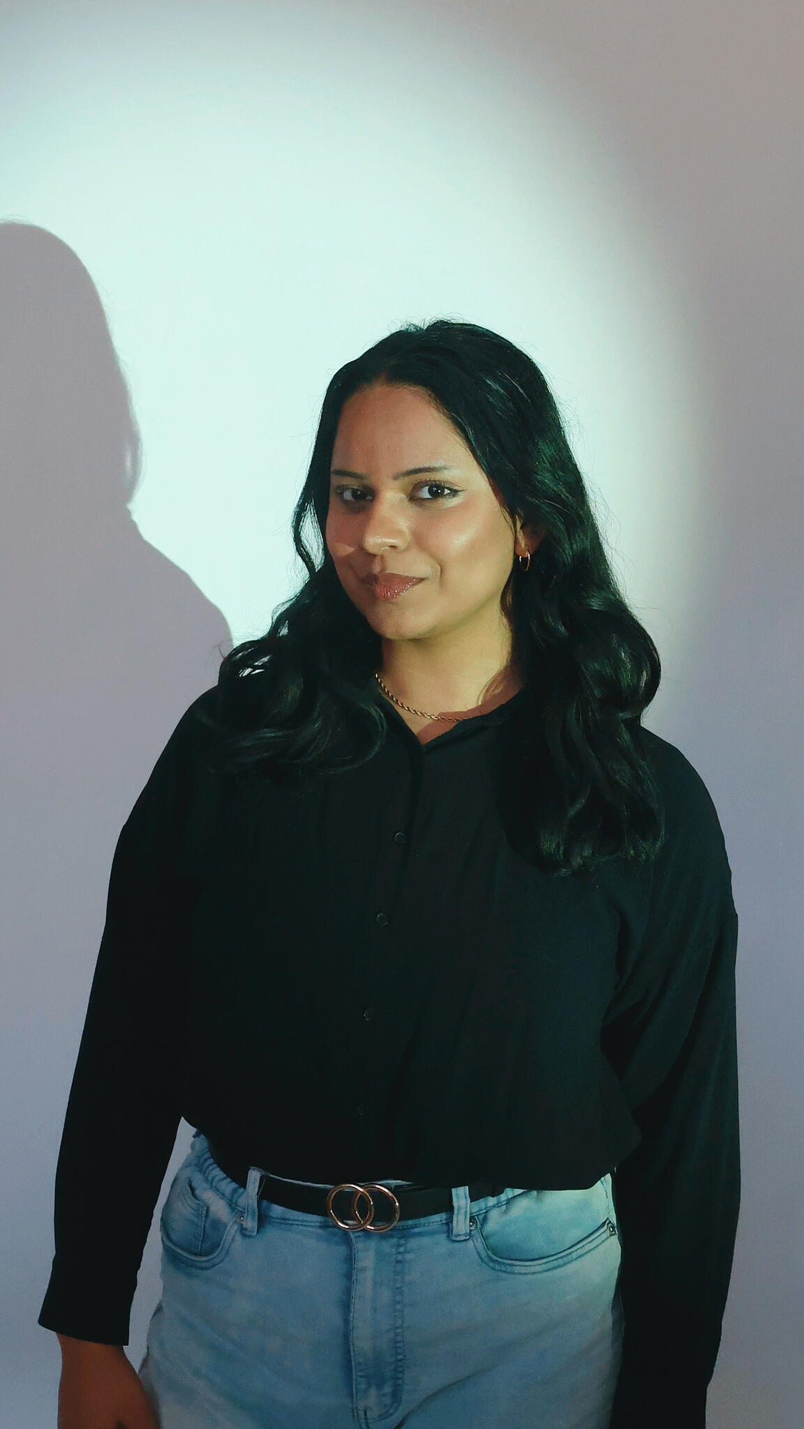 Kristen Joanes stands in front of a white wall with lighting highlighting her face. She wears a black shirt and jeans and smiles standing with her head angled.