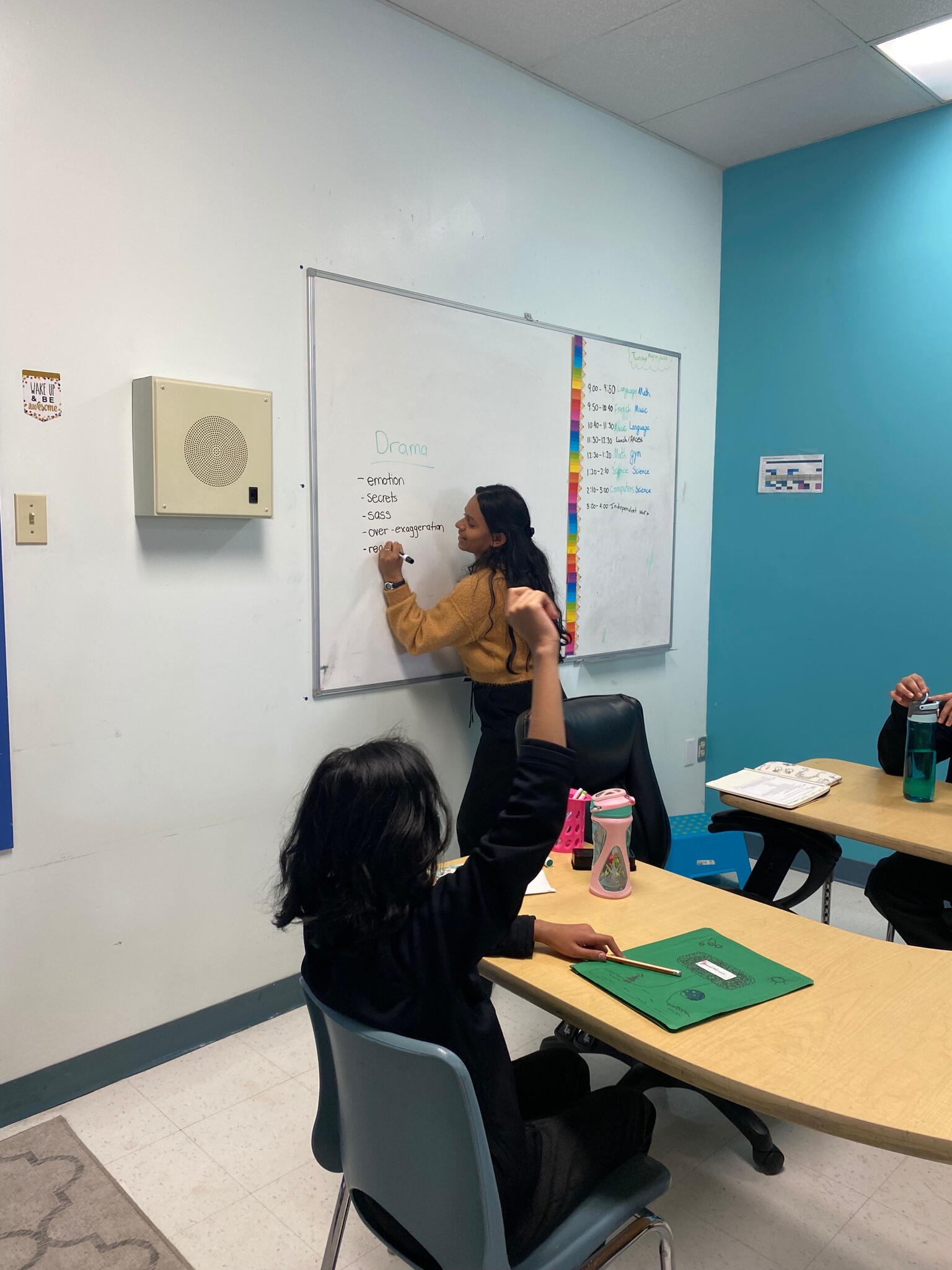 Kristen stands a white board and writes with a black marker while smiling. A student in the foreground raises their hand.
