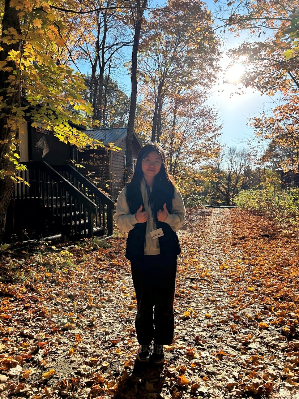 Wennie stands on a path covered in fallen fall leaves. She wears a vest and there is sun shining in the background along with the deck of some log cabins.