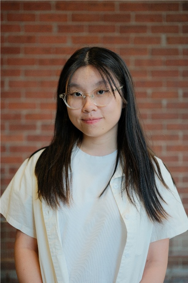 Wennie Chen smiles wearing a white shirt and white button down t-shirt over top and glasses. Her hair is down around her shoulders and she stands in front of a red brick wall.