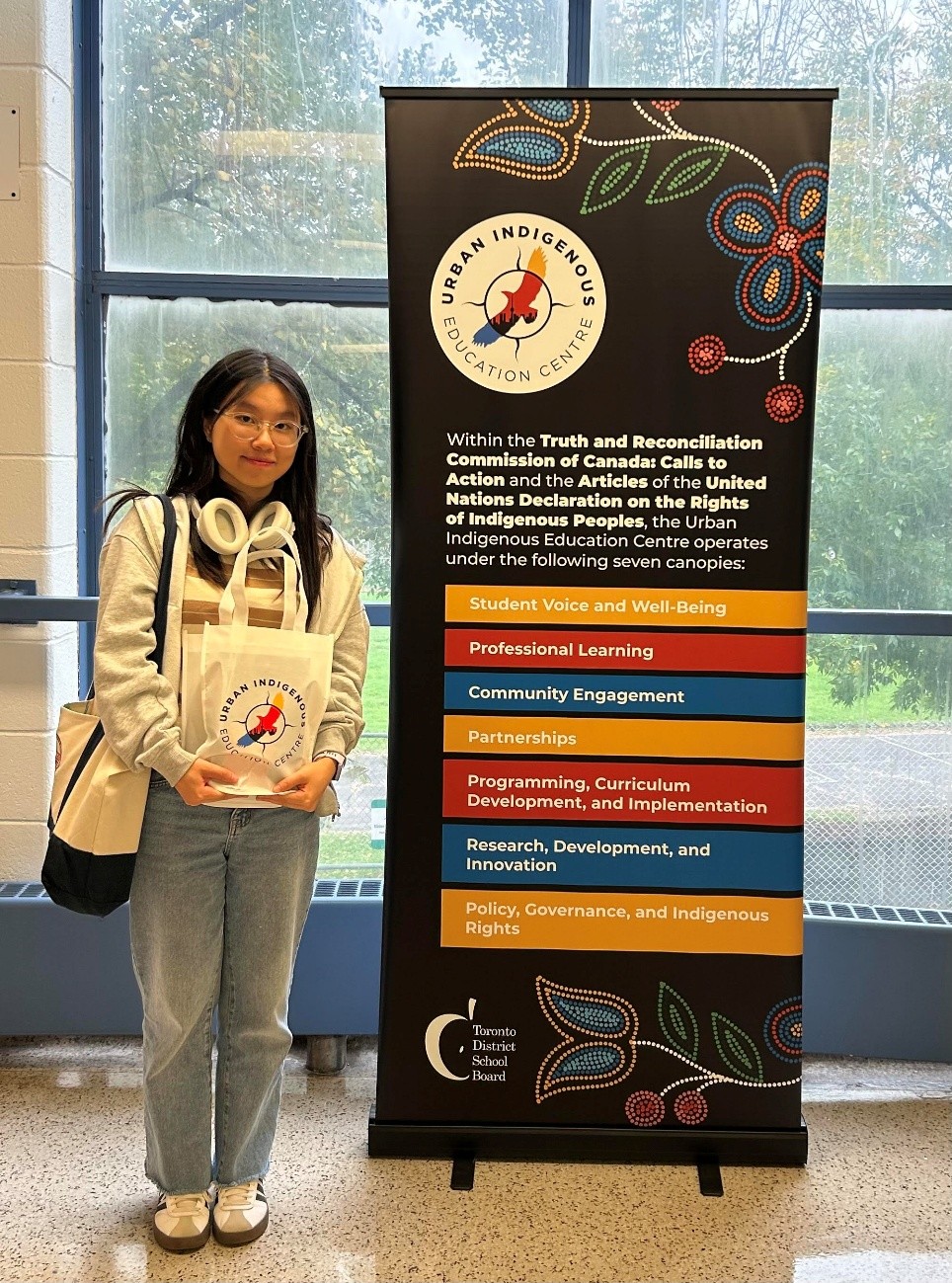 Wennie holds a bag and stands in a school hallway beside a standing banner that reads "Urban Indigenous Education Centre" and explains the TDSB's seven canopies.