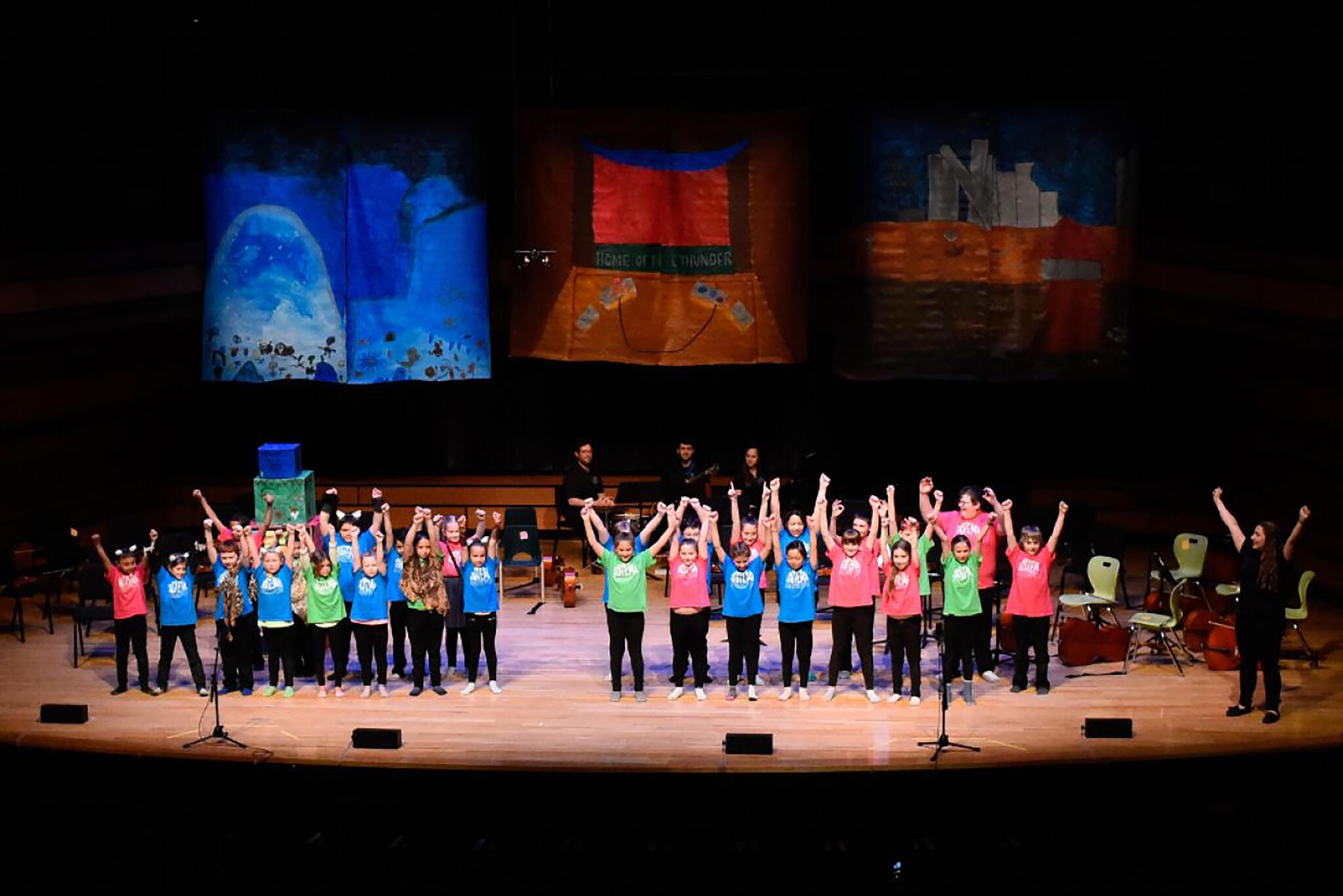 Children in colourful t-shirts stand on a stage with their arms raised preparing to bow.