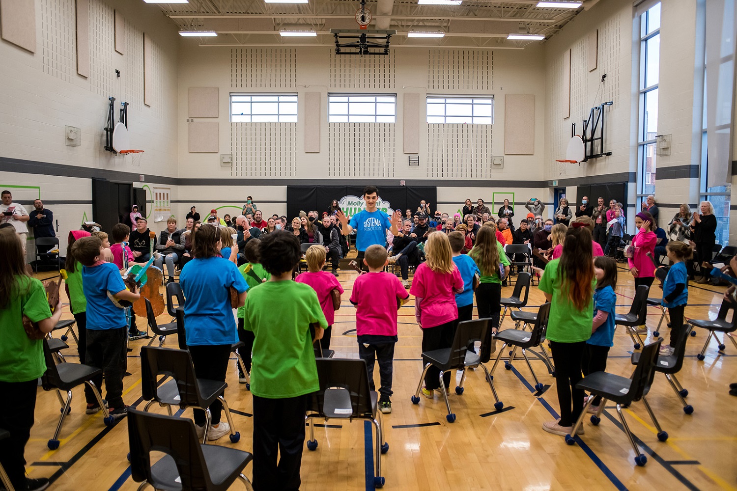 A view of Sistema Kingston students performing at a concert. Their backs face the camera and they face their parents sitting in the audience.
