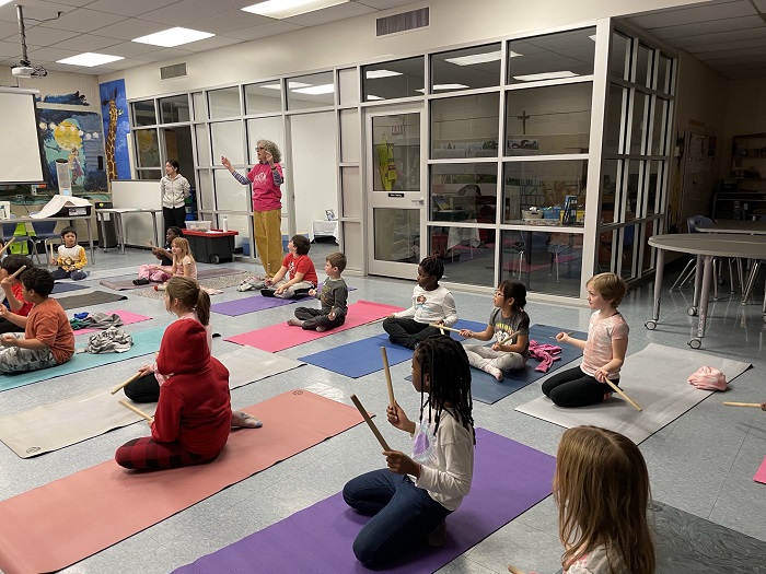 A teacher raises her arms to instruct students that hold on to rhythm sticks while seated on the ground.