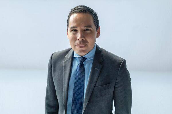 Natan Obed smiles in front of a grey background wearing a suit.