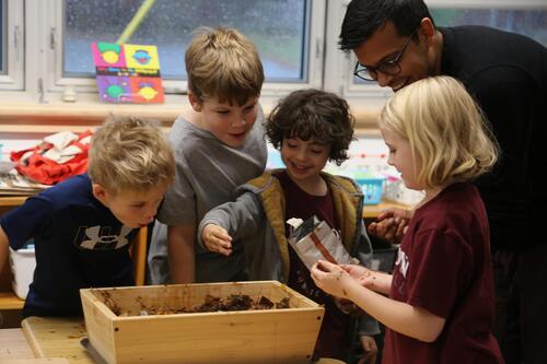 Students reach into a wooden box filled with dirt while a teacher supervises