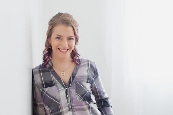 Lindsay Carmichael smiles leaning against a white wall in a plaid purple shirt.