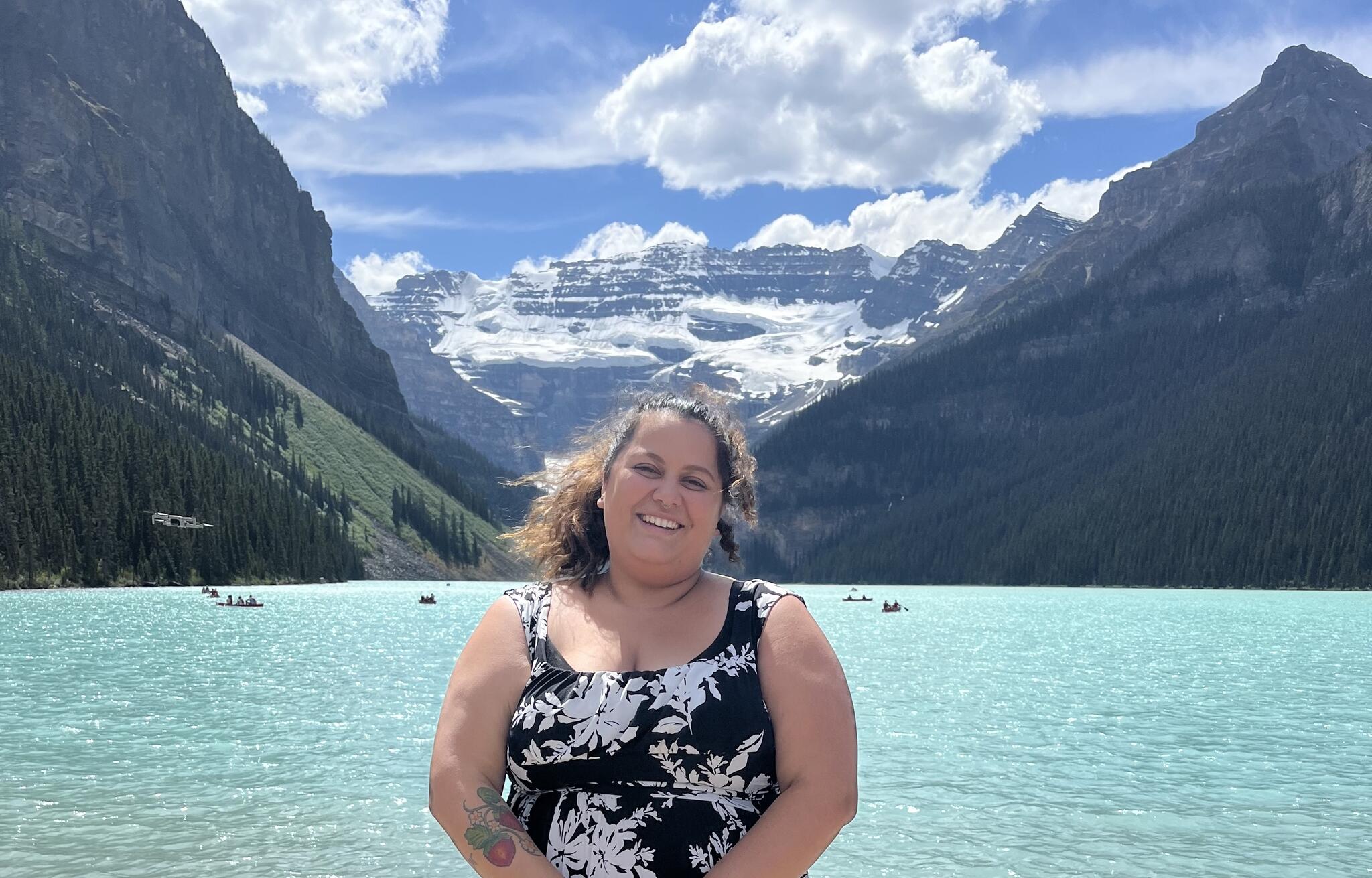 A woman in a black and white floral dress smiles at the camera in front of crystal blue water and tall mountains
