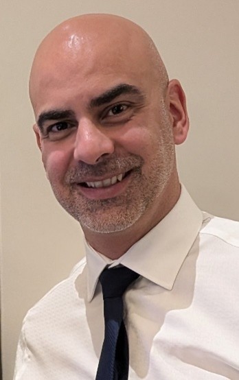 A man with bald hair smiles at a camera wearing a navy tie and white shirt