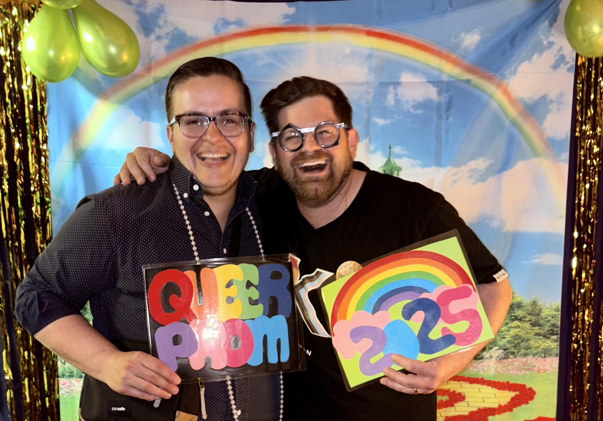 Two people smiling with signs that say Queer Prom 2025 and a rainbow behind them