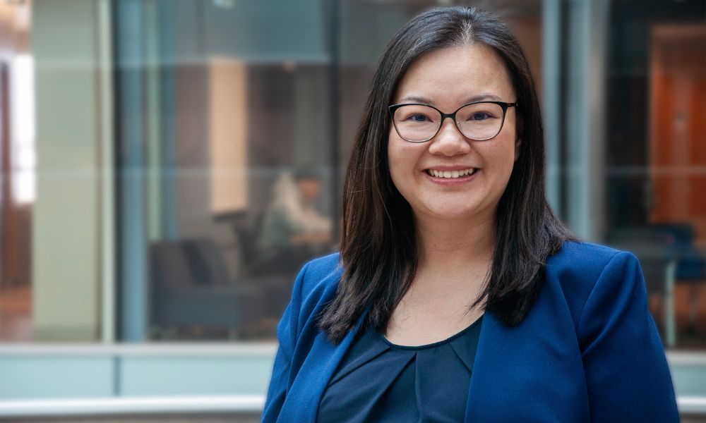 An Asian Woman wearing glasses smiles with a blue blazer on 