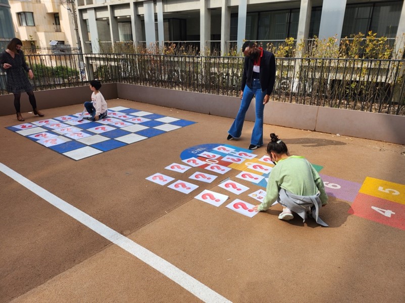 A woman black woman stands on a playground with her head tilted to one side looking at two children working on an exercise with pieces of paper with large question marks on them. 