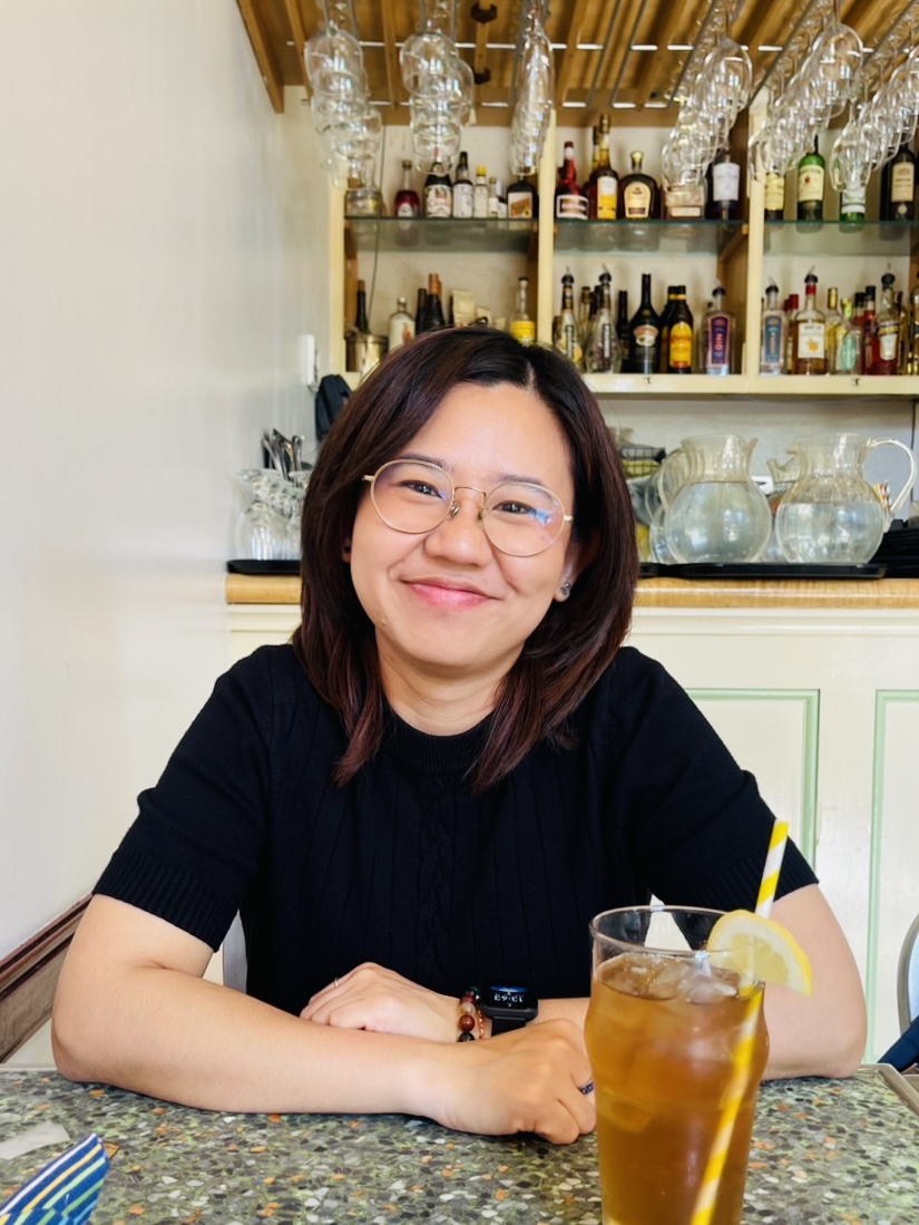 A woman with dark hair smiles at the camera at a restaurant with iced tea on the table