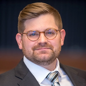 A white man with glasses and wearing a suit looks straight into the camera 