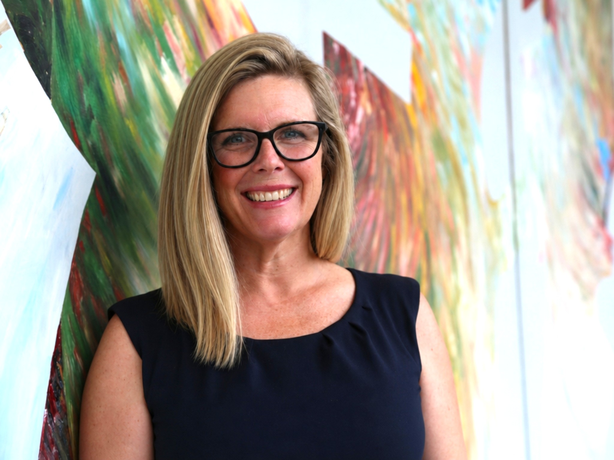 A woman smiles at the camera with blond hair and wearing glasses and posing in front of art 