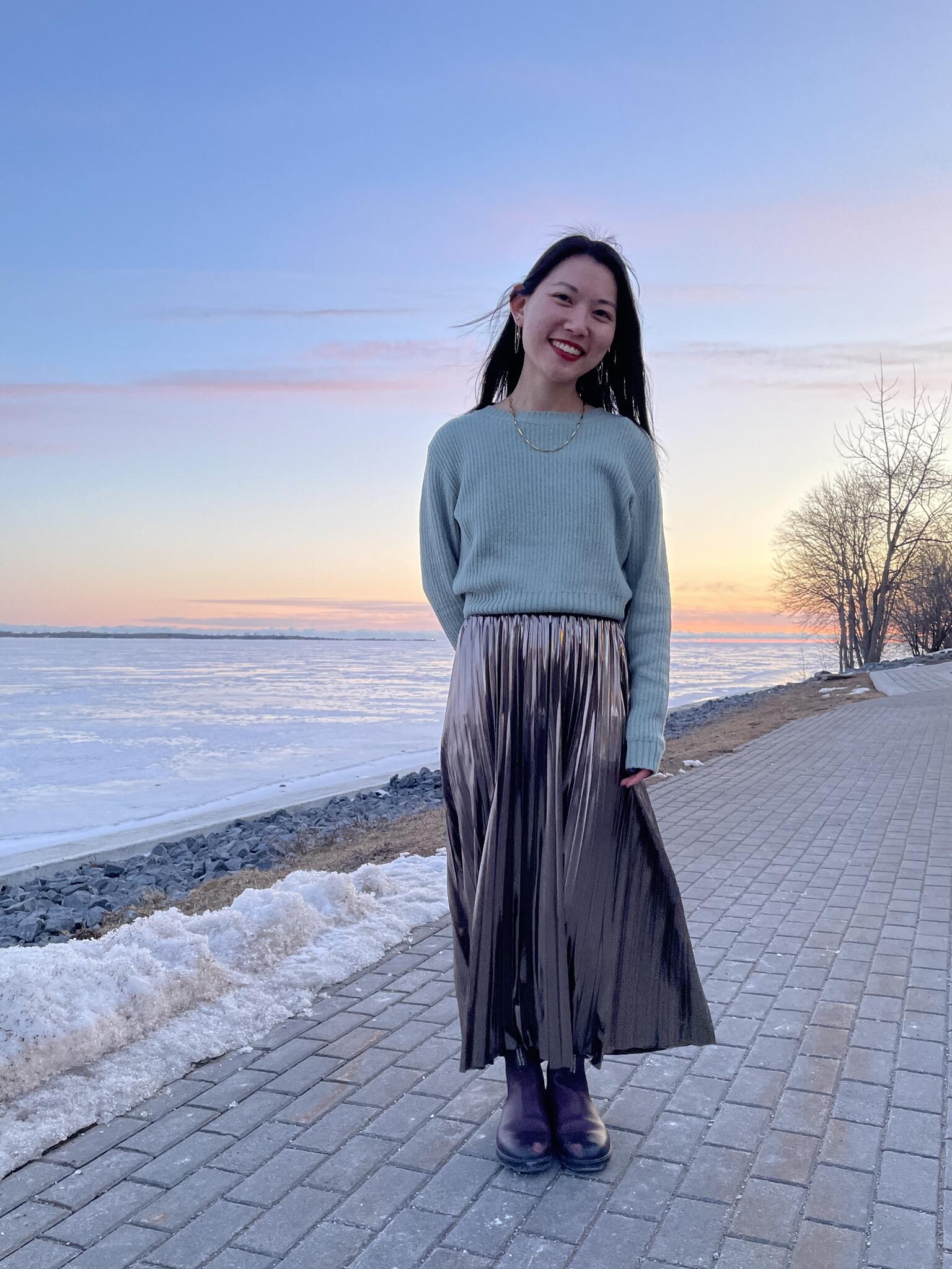 An Asian woman posing on a water front in a blue sweater and metallic long skirt in the winter at sunset