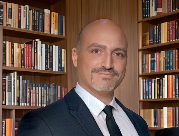 A man with a bald head wearing a suit and a tie posing in front of book shelves