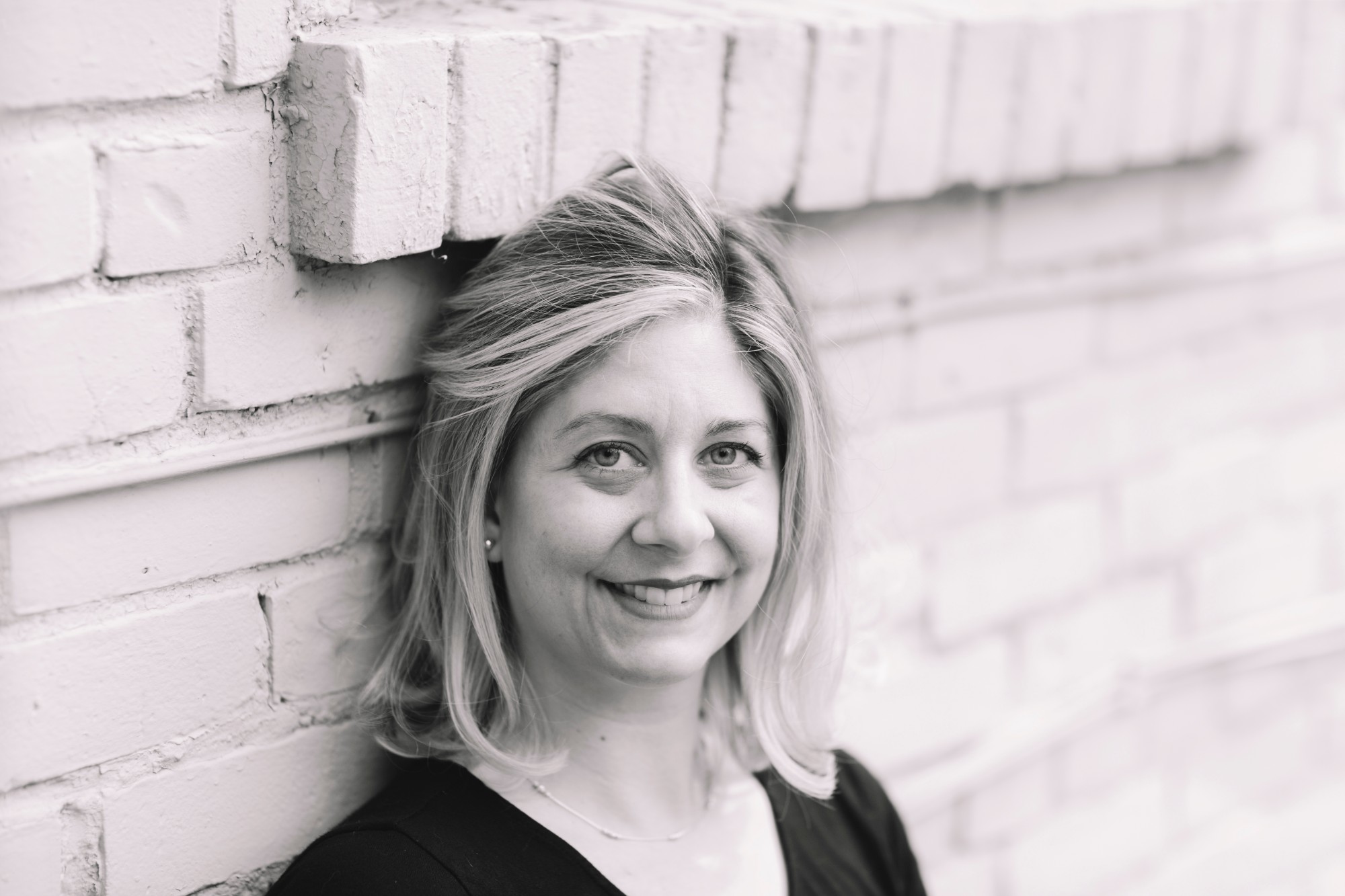 A white blonde woman smiles at the camera in front of a white brick building