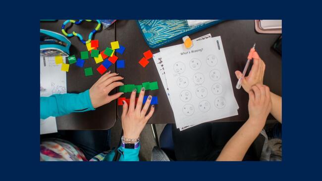 A bird's eye view of students working on a math sheet with colourful squares on the table.