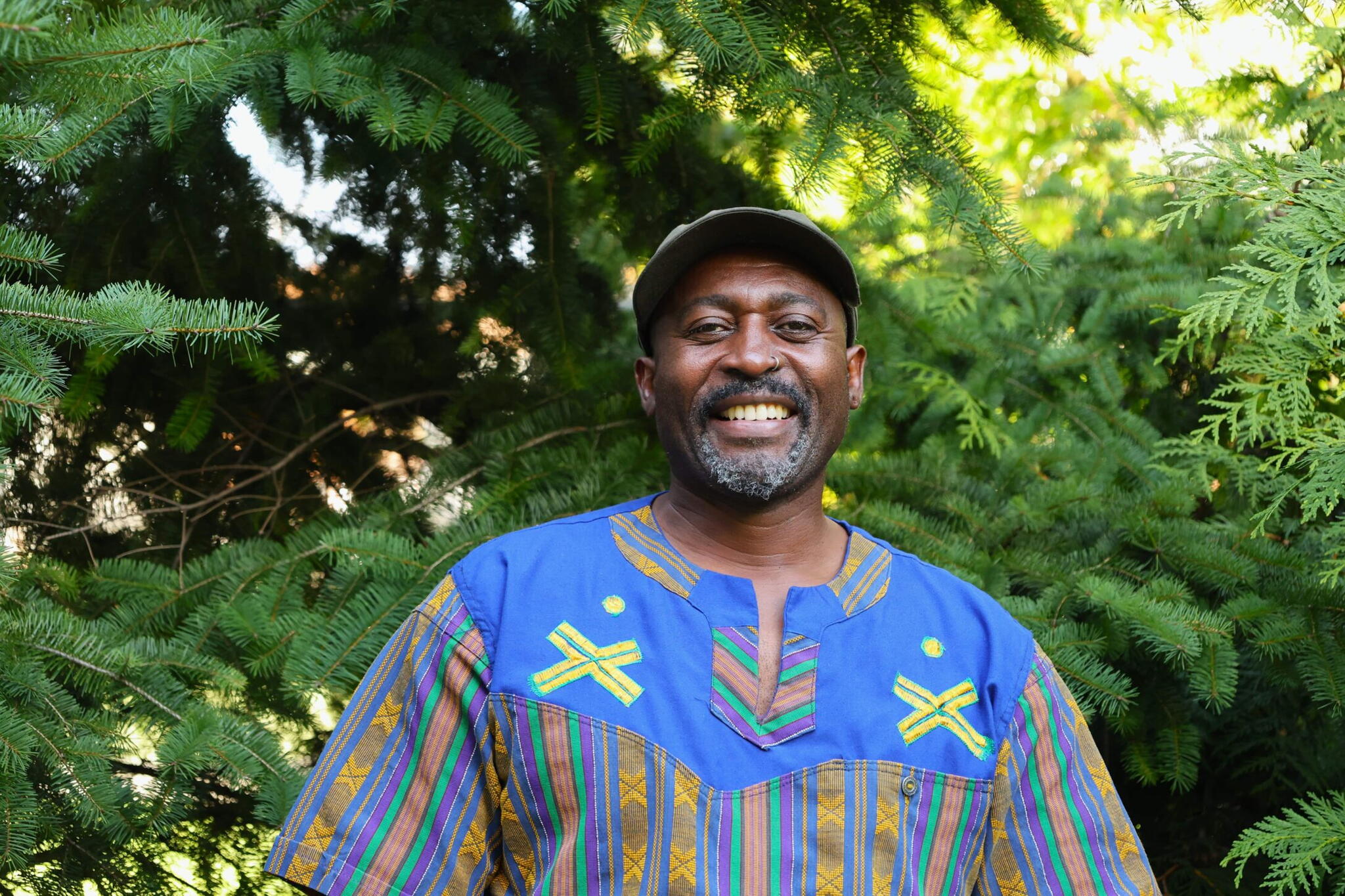 Dr. Devi Mucina smiles wearing a patterned shirt and hat. He stands in front of a group of lush green trees.