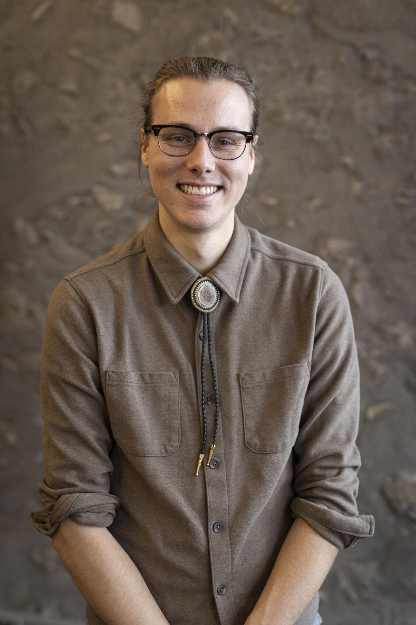 Jackson Pind smiles wearing a brown button down shirt and bolo tie. There is a mottled brown background behind Jackson.