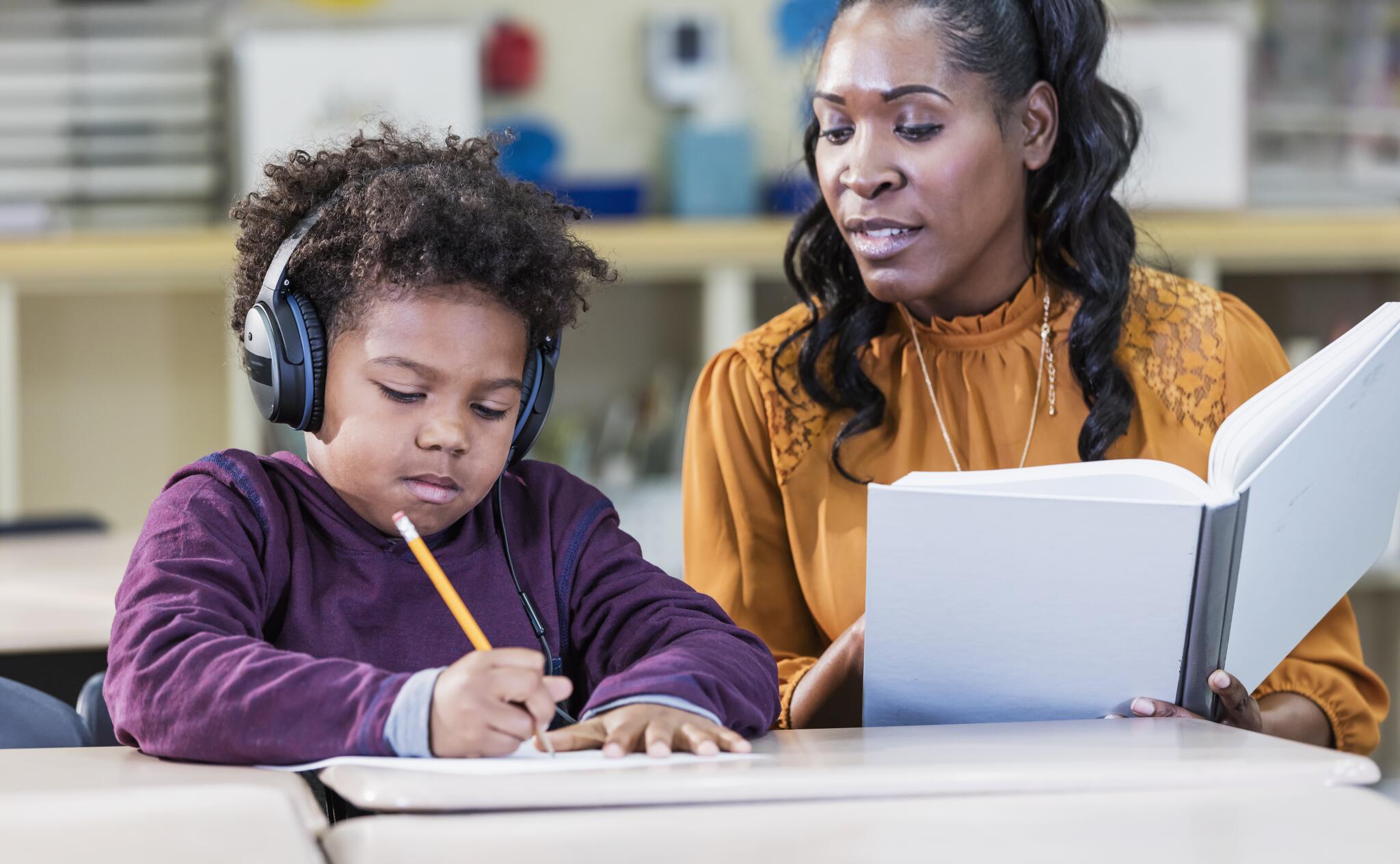 A teacher works with a student wearing headphones and holding a pencil.
